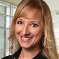Smiling researcher with long blonde hair, wearing a black jacket, poses in a bright, modern setting.